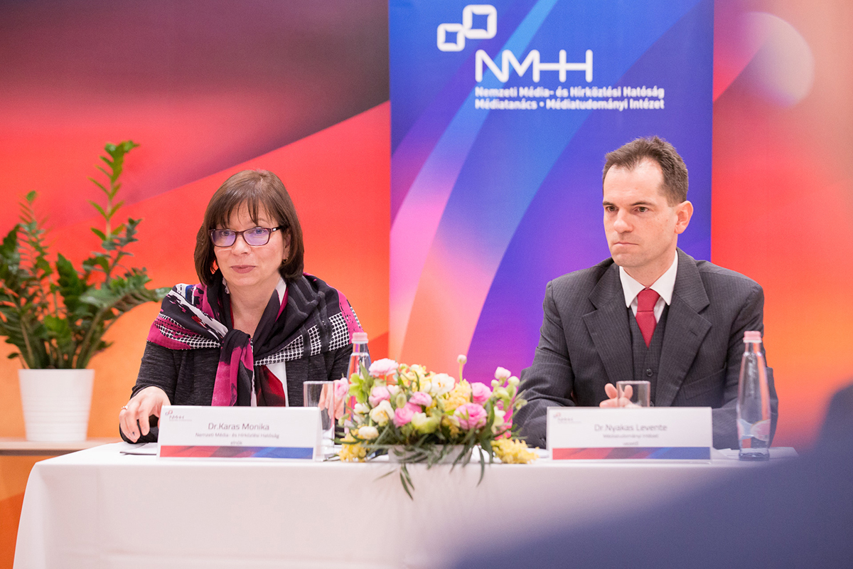 A kép bal oldalán dr. Karas Monika, az NMHH elnöke, jobbra dr. Nyakas Levente, az NMHH Médiatanács Médiatudományi Intézet vezetője látható az elnöki asztalnál. Karas Monika éppen beszél a jelen lévő vendégekhez