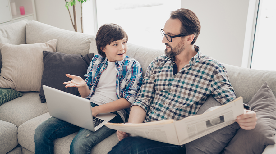 Egy 13-14 éves forma gyermek laptoppal az ölében ül egy kanapén az újságot olvasó édesapja melett, és éppen beszélgetnek.